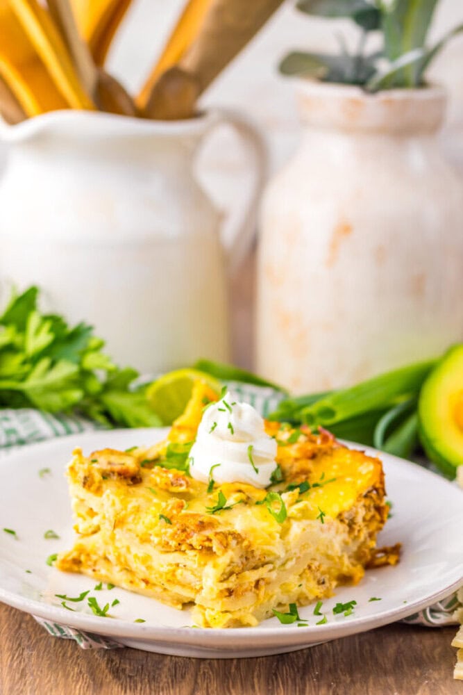 A slice of egg and cheese casserole topped with sour cream and chopped herbs is served on a white plate, with avocado and fresh greens in the background.