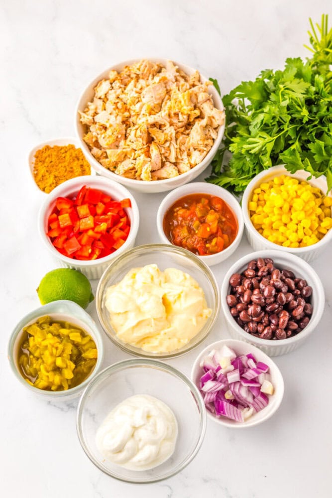 A variety of ingredients in bowls, including shredded chicken, mayonnaise, sour cream, black beans, salsa, red onion, corn, red pepper, pickled jalapeños, lime, and fresh parsley.
