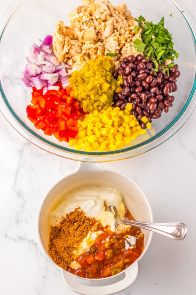 A glass bowl with chopped chicken, red onion, parsley, diced pickles, black beans, red bell pepper, and corn; below, a bowl with mayonnaise, sour cream, salsa, and taco seasoning.