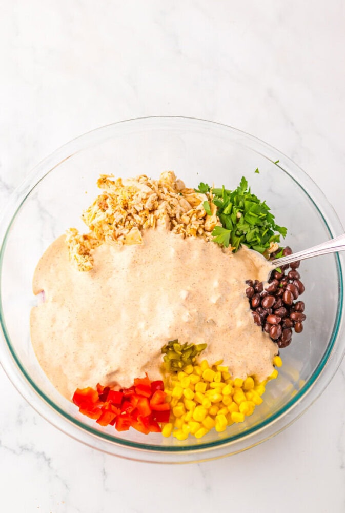 A glass bowl contains shredded chicken, chopped cilantro, black beans, corn, red bell pepper, jalapeños, and a creamy dressing, with a spoon resting on the side.