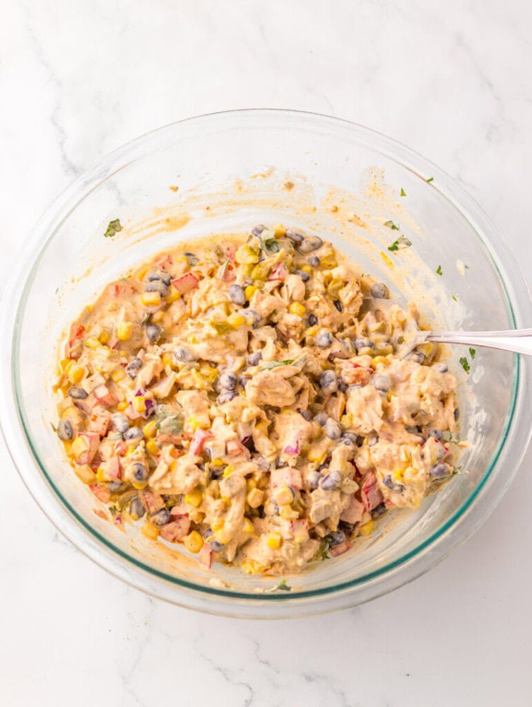 A glass bowl filled with a mixture of chopped vegetables, black beans, corn, and creamy dressing, with a spoon resting inside.
