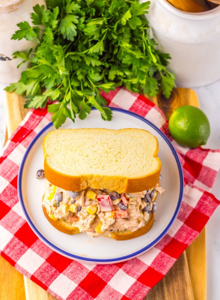 Chicken salad sandwich with vegetables on white bread, served on a plate with a red checkered cloth, parsley, and a lime in the background.