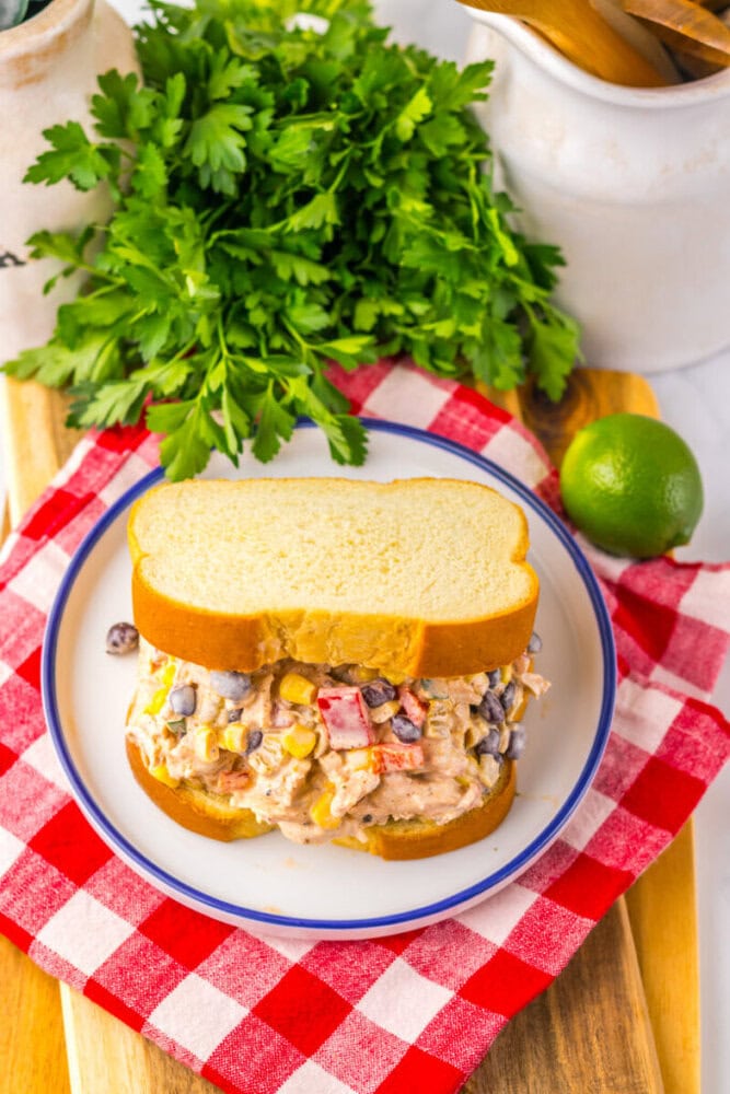A chicken salad sandwich with beans and vegetables on white bread sits on a white plate, with fresh parsley and a lime in the background on a red checkered cloth.