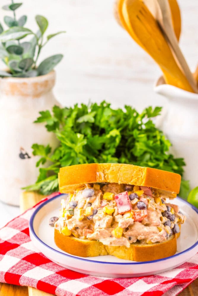 A chicken salad sandwich with corn, black beans, and diced peppers on white bread sits on a plate, with parsley and kitchen utensils in the background.