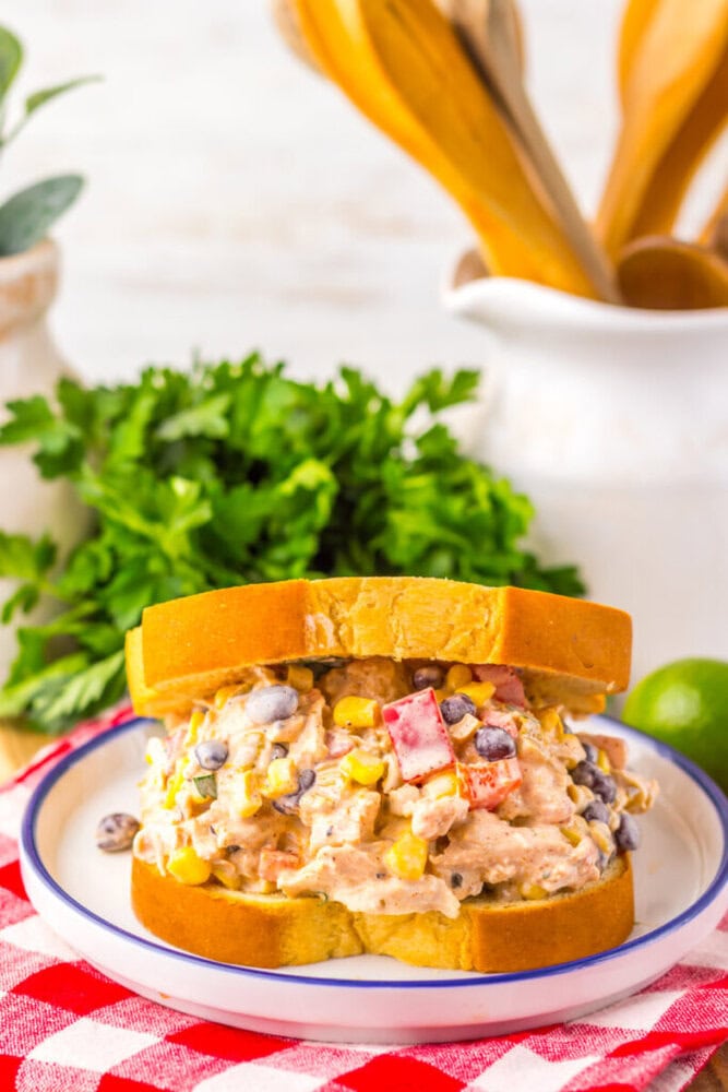 A chicken salad sandwich with corn, black beans, and diced red peppers on yellow bread, served on a white plate with a red-checkered napkin. Parsley and kitchen utensils in the background.