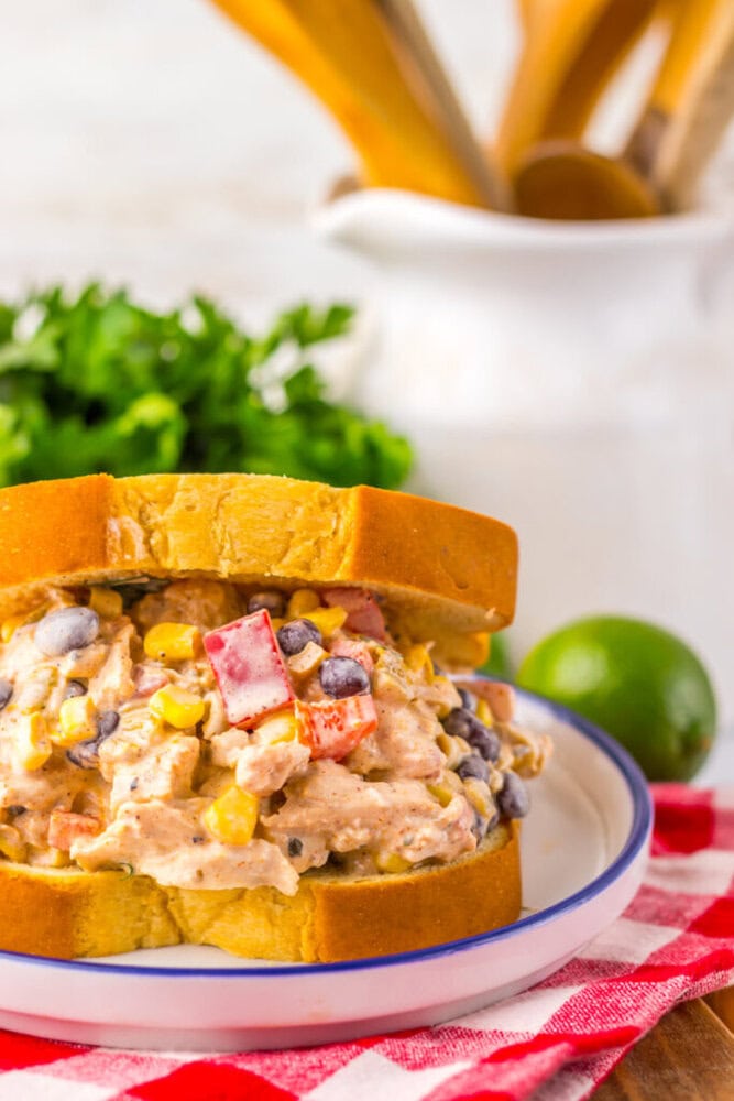 A close-up of a chicken salad sandwich with corn, black beans, and diced red peppers on a white plate, set on a red checkered cloth.