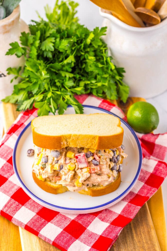 A chicken salad sandwich with mixed vegetables on white bread sits on a plate, with fresh parsley, a lime, and a red checkered napkin nearby.