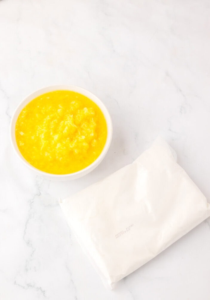 A white bowl filled with beaten eggs sits next to a sealed packet on a white marble surface.