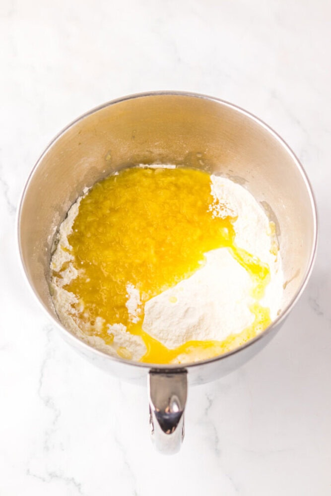 A metal mixing bowl containing flour and melted butter on a white marble surface, viewed from above.