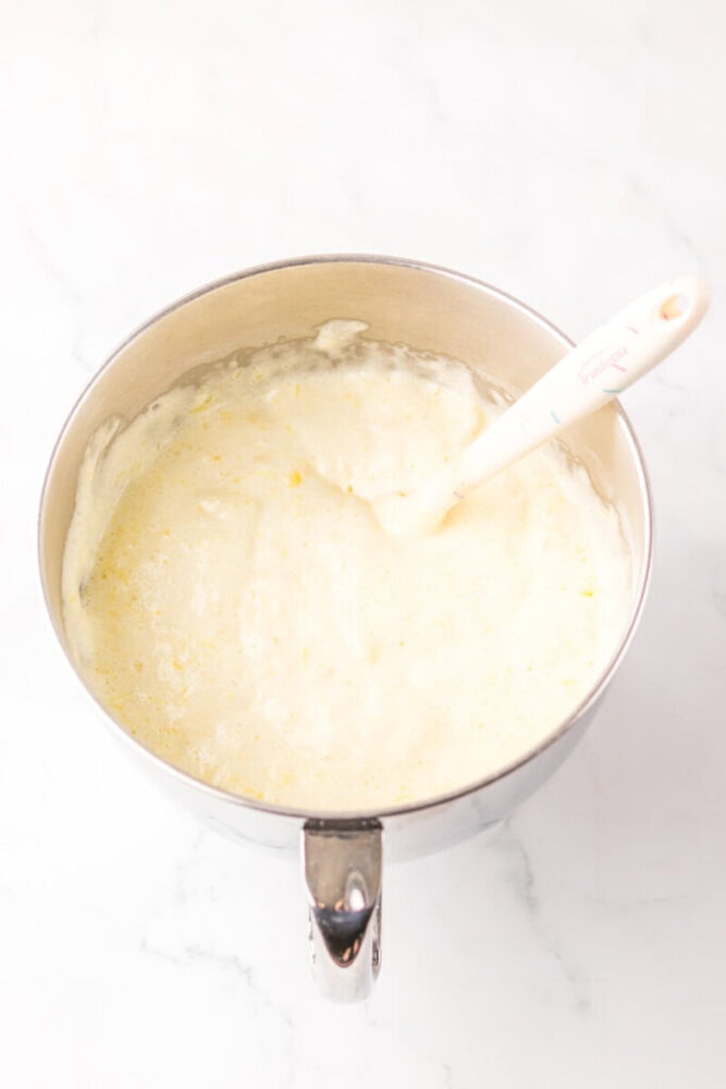 A mixing bowl filled with a pale, foamy batter being stirred with a white spatula on a white countertop.