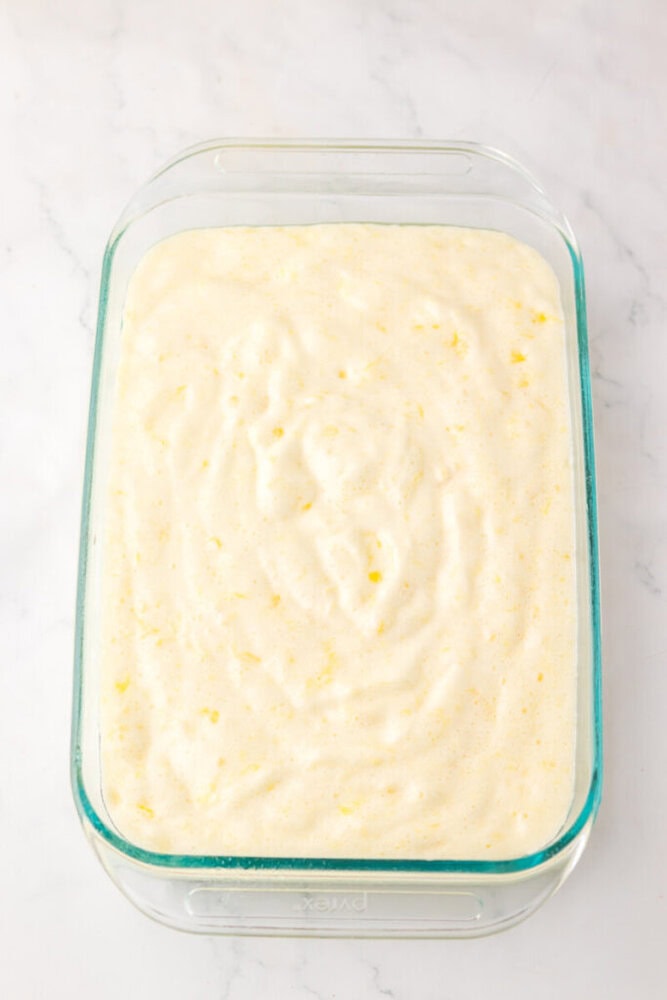A glass baking dish filled with an unbaked, light yellow, fluffy batter on a white marble surface.