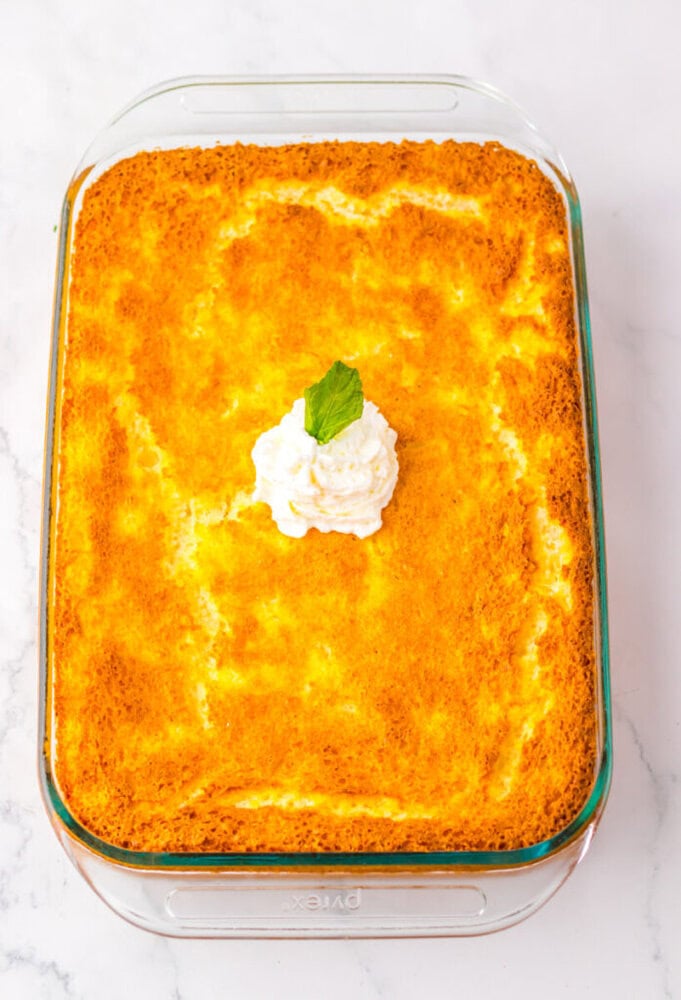 A baked yellow cake topped with a dollop of whipped cream and a mint leaf, served in a rectangular glass dish on a white surface.