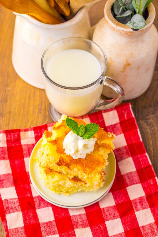 A plate with two pieces of cornbread topped with whipped cream and mint, next to a glass mug of milk on a red and white checkered napkin.