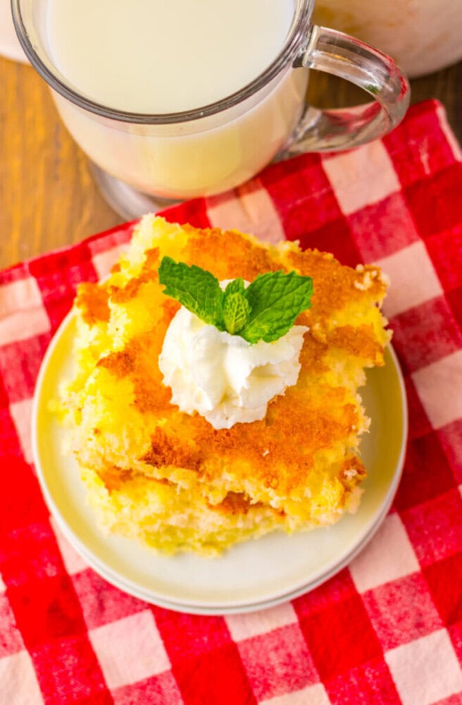 A slice of yellow cake topped with whipped cream and a mint leaf on a plate, next to a glass of milk, set on a red and white checkered cloth.