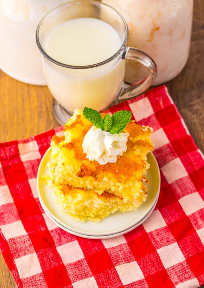 A plate with two pieces of yellow cake topped with whipped cream and mint sits on a red checkered cloth next to a glass mug of milk.