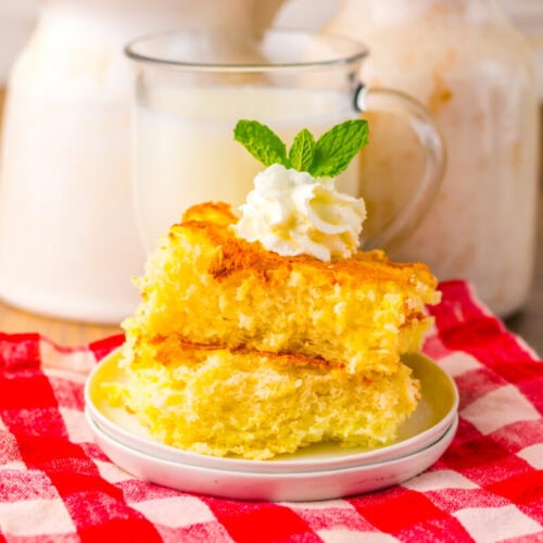 Two slices of yellow cake topped with whipped cream and a mint leaf on a small plate, placed on a red checkered cloth, with a glass of milk in the background.