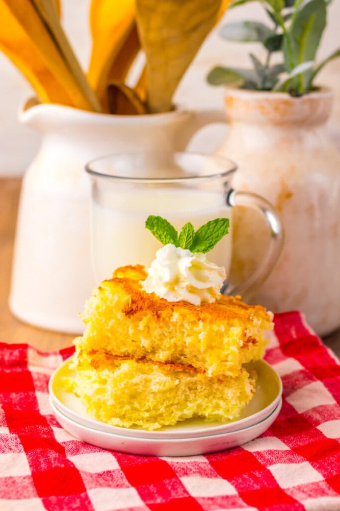Two pieces of yellow cake topped with whipped cream and a mint sprig on a plate, with a glass of milk and decorative jars in the background.