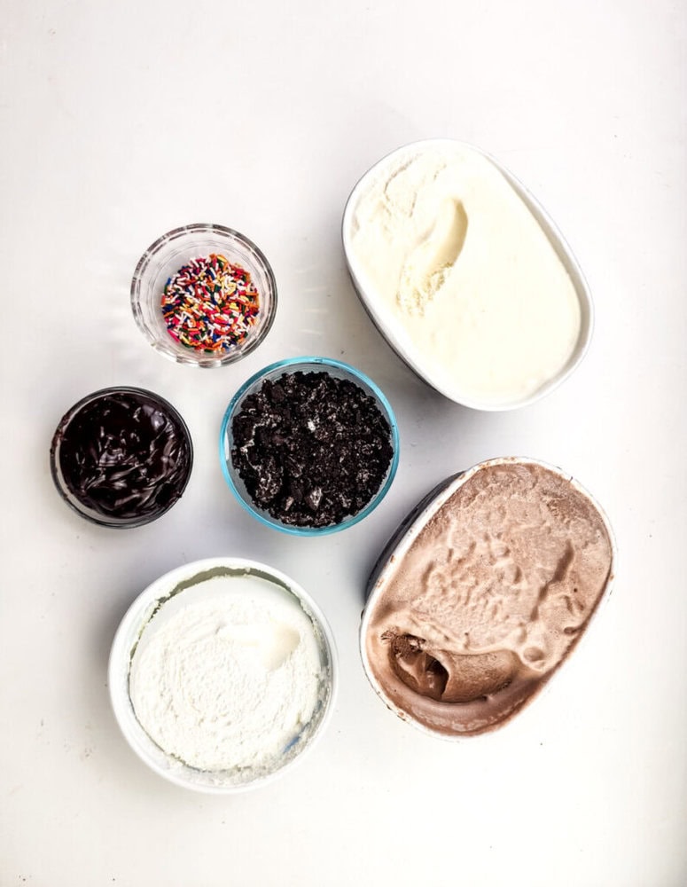 Bowls with vanilla and chocolate ice cream, whipped cream, chocolate syrup, crushed cookies, and colorful sprinkles arranged on a white surface.