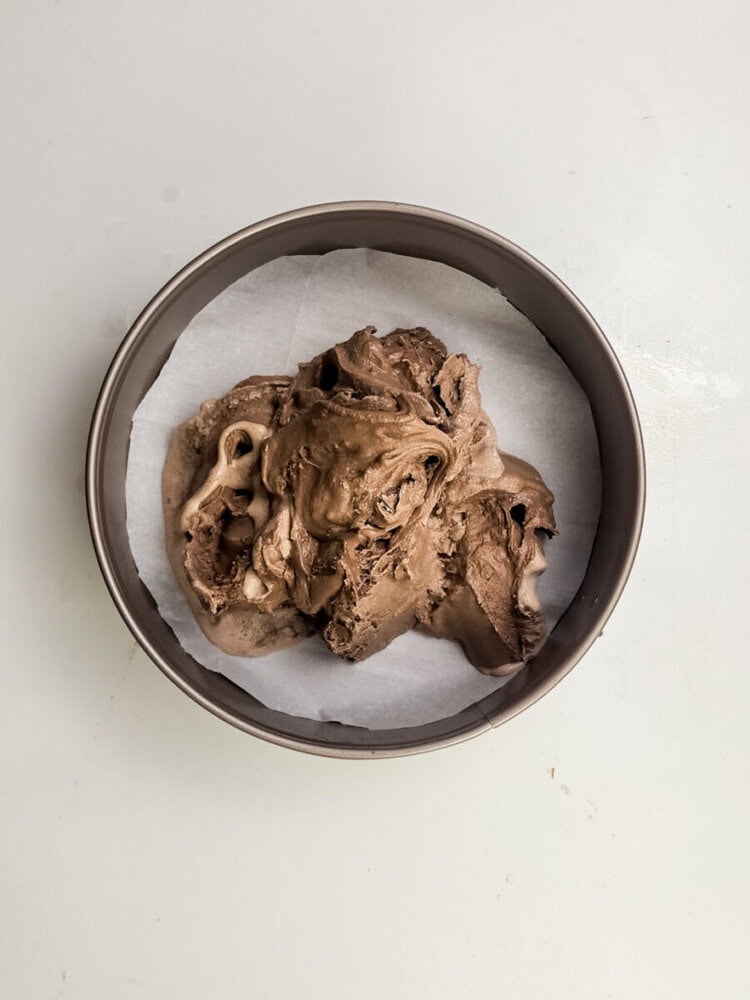 Chocolate ice cream is spread on a round layer of parchment paper in a metal cake pan, viewed from above on a white surface.