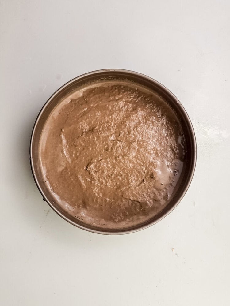 A round cake pan filled with unbaked chocolate cake batter on a plain white surface.