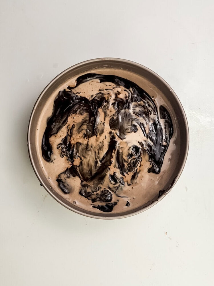 A round pan filled with chocolate batter, swirled with a dark chocolate mixture, sits on a white surface.
