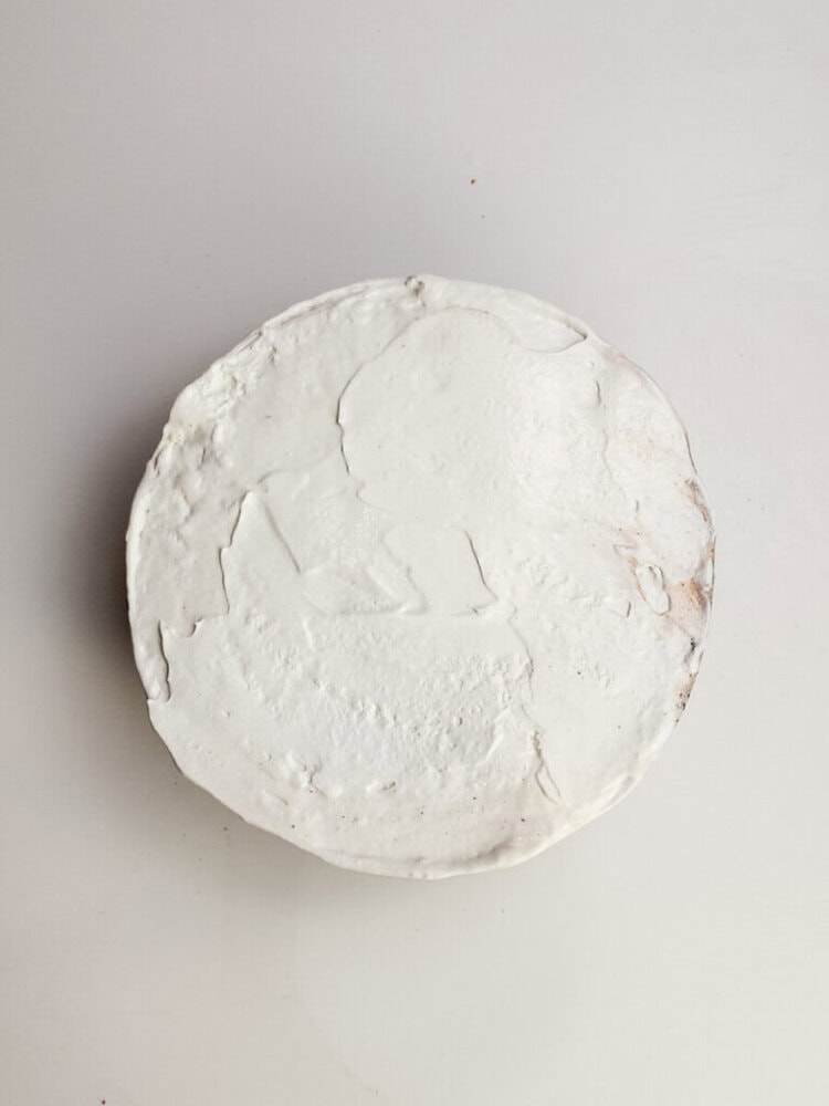 A round white plaster or clay disc with an uneven surface and faint, indiscernible impressions, placed on a plain white background.