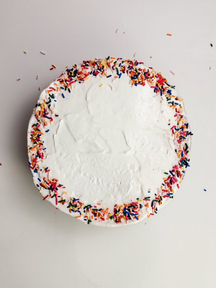 A round cake with white frosting, decorated with rainbow sprinkles around the edge, sits on a white surface.