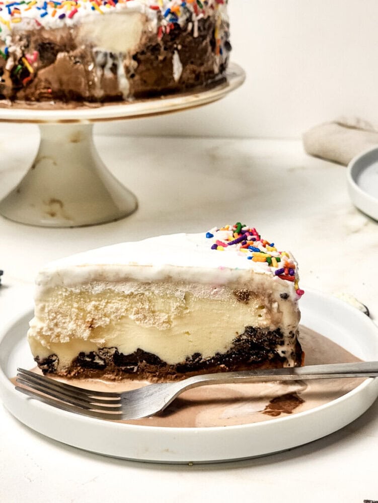 A slice of layered ice cream cake with chocolate base, vanilla ice cream, whipped topping, and rainbow sprinkles on a white plate with a fork. The whole cake is in the background.