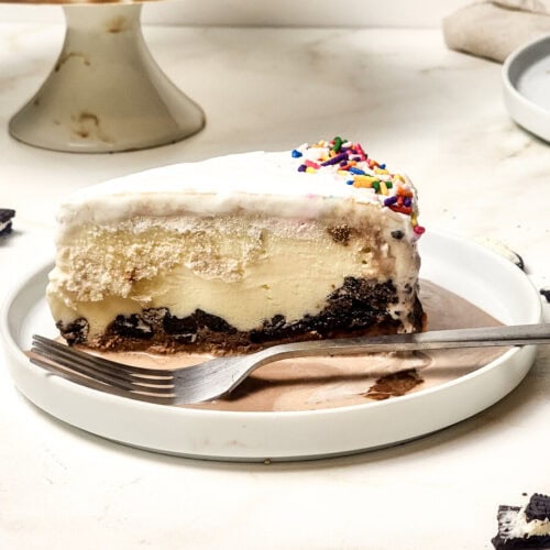 A slice of ice cream cake with a chocolate base, vanilla layers, white frosting, and rainbow sprinkles, served on a plate with a fork. Broken cookies and another plate are nearby.