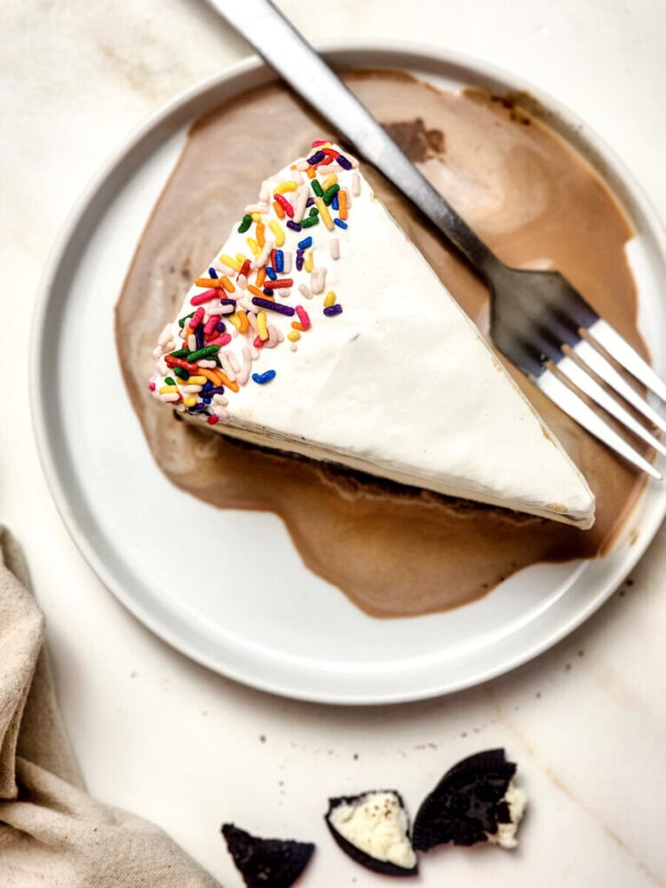A slice of cake with white frosting and rainbow sprinkles on a white plate, with a fork and some chocolate sauce, next to broken Oreo cookies.