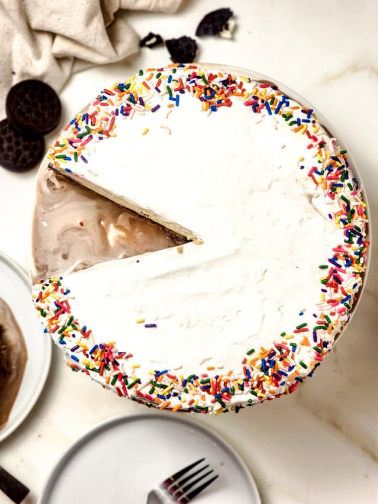 A round ice cream cake with a whipped topping and rainbow sprinkles along the edge, one slice removed and placed on a nearby plate.