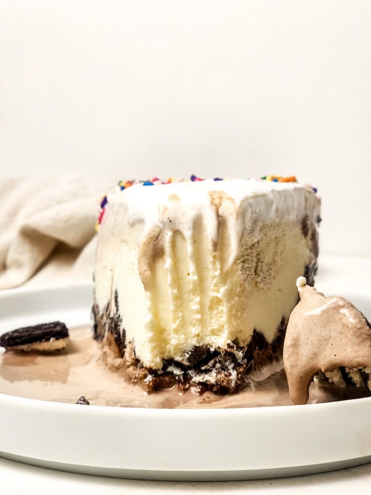 A partially eaten slice of ice cream cake with chocolate layers and rainbow sprinkles on a white plate, next to a spoonful of melted ice cream.