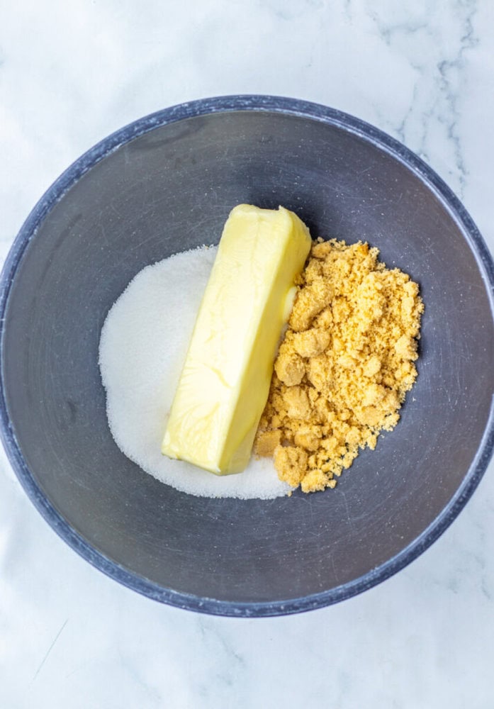 A mixing bowl containing a stick of butter, granulated sugar, and brown sugar on a white countertop.