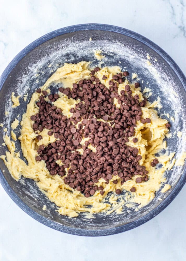 A mixing bowl filled with cookie dough and topped with a large amount of chocolate chips, ready to be mixed.