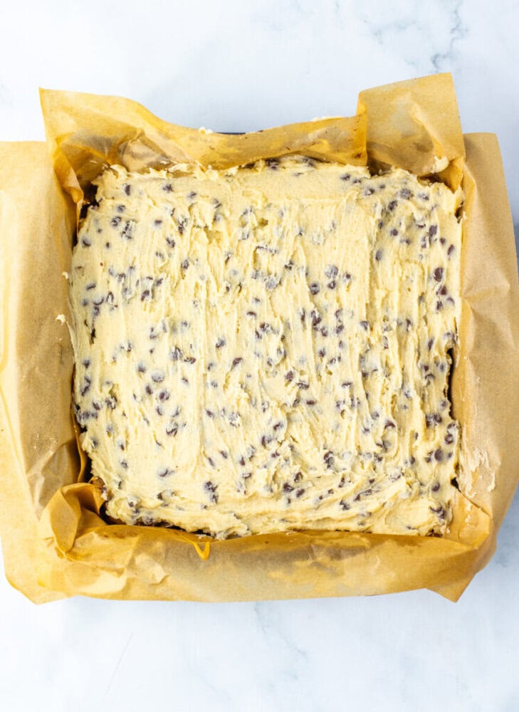 A square baking pan lined with parchment paper is filled with unbaked chocolate chip cookie dough, spread evenly.