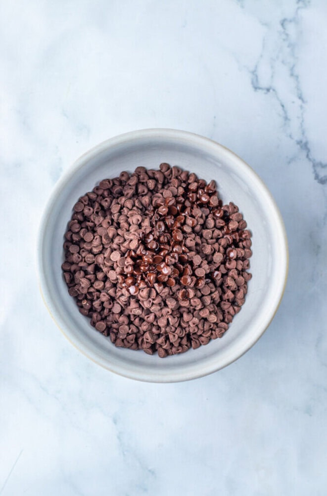 A white bowl filled with semi-sweet chocolate chips, some of which are partially melted, on a light marble surface.