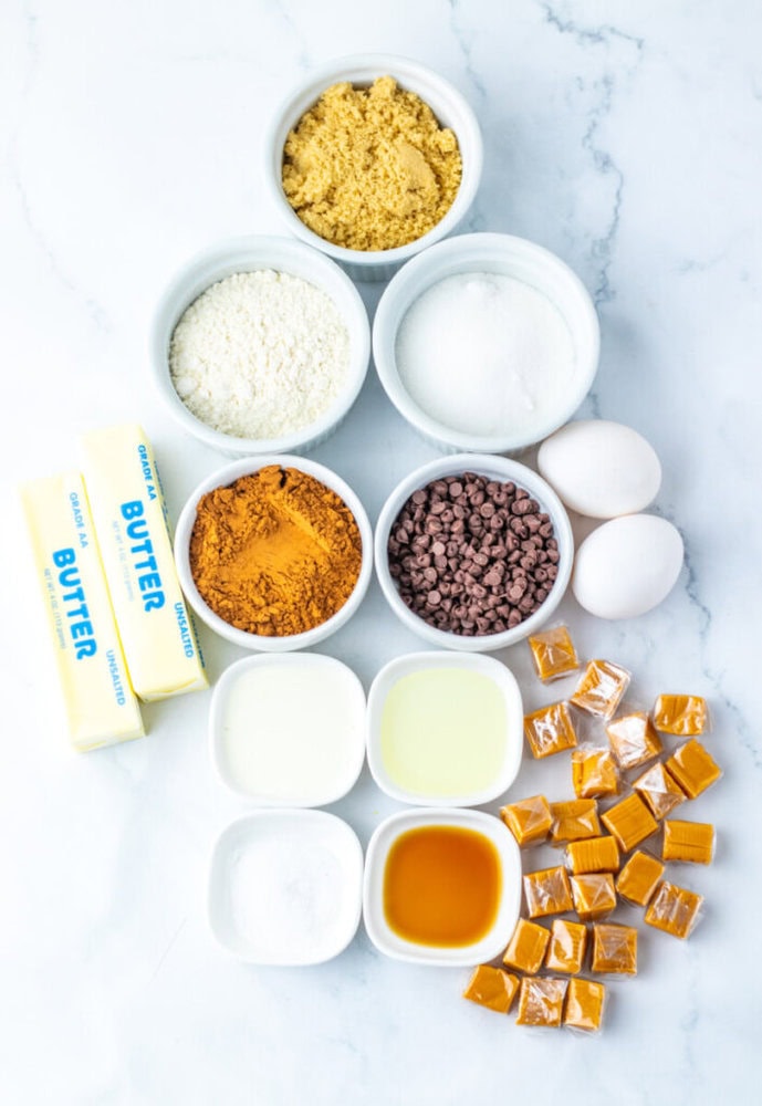 Various baking ingredients including butter, brown sugar, flour, granulated sugar, eggs, cocoa powder, chocolate chips, caramel candies, baking powder, oil, milk, and vanilla extract.