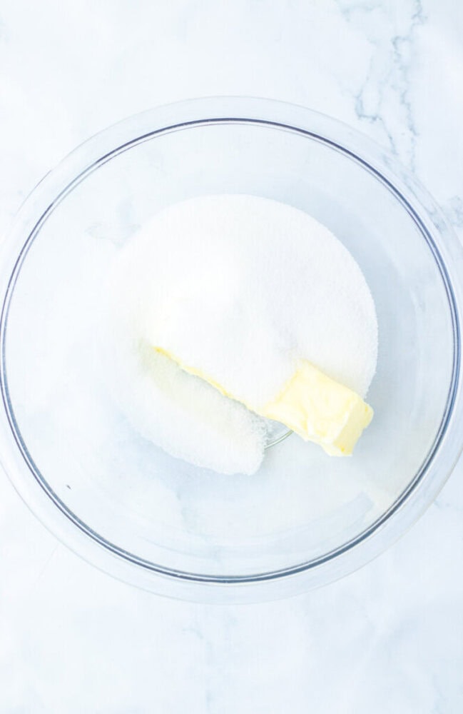 A clear glass bowl containing a stick of butter and a pile of granulated white sugar on a white marble surface.