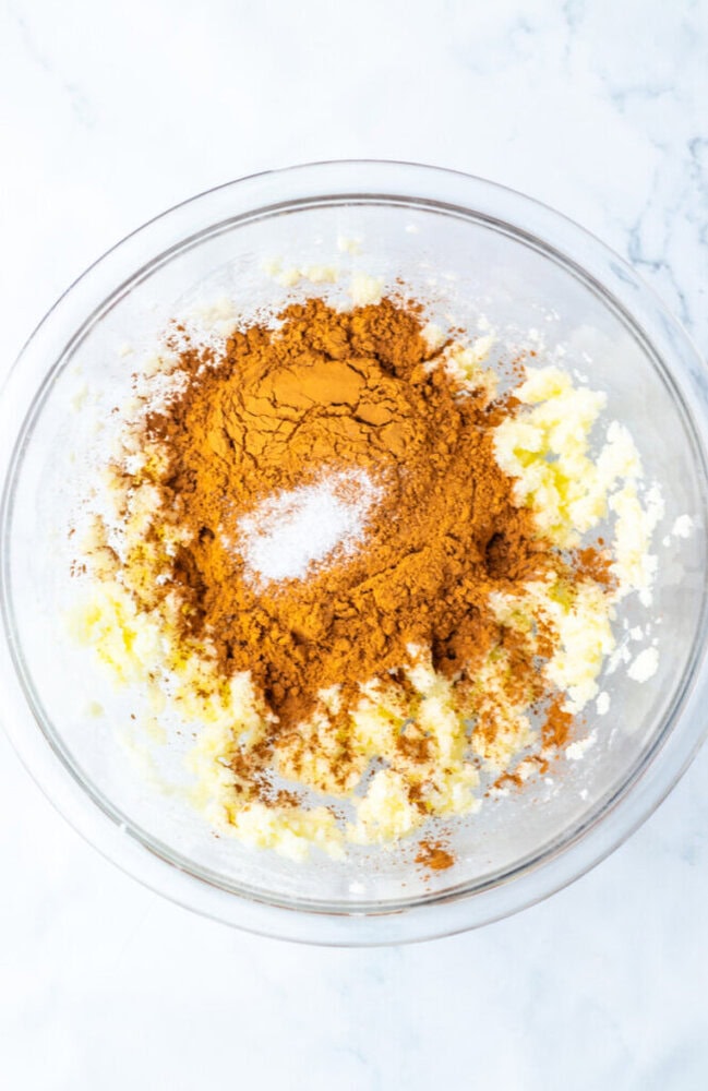 A glass mixing bowl containing creamed butter and sugar with ground cinnamon and a small amount of salt on top, viewed from above on a white surface.