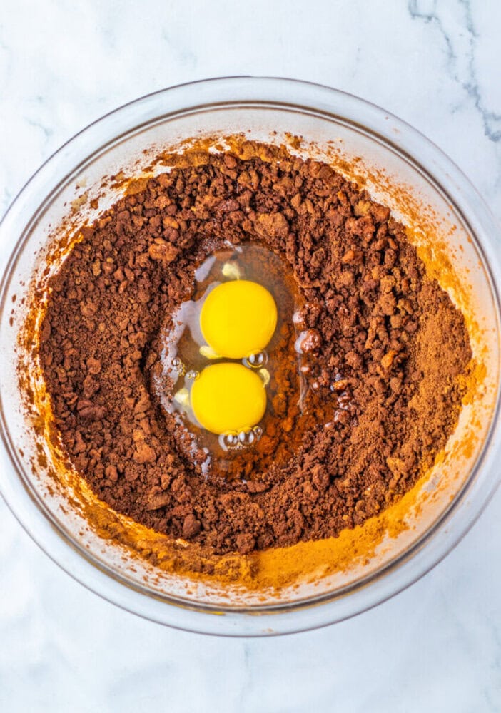 A glass mixing bowl containing cocoa powder and two raw eggs on a marble surface.