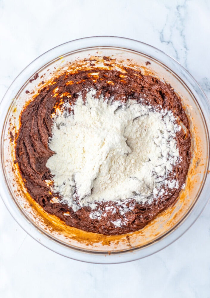 A glass bowl containing chocolate batter with a mound of white flour on top, placed on a white marble surface.