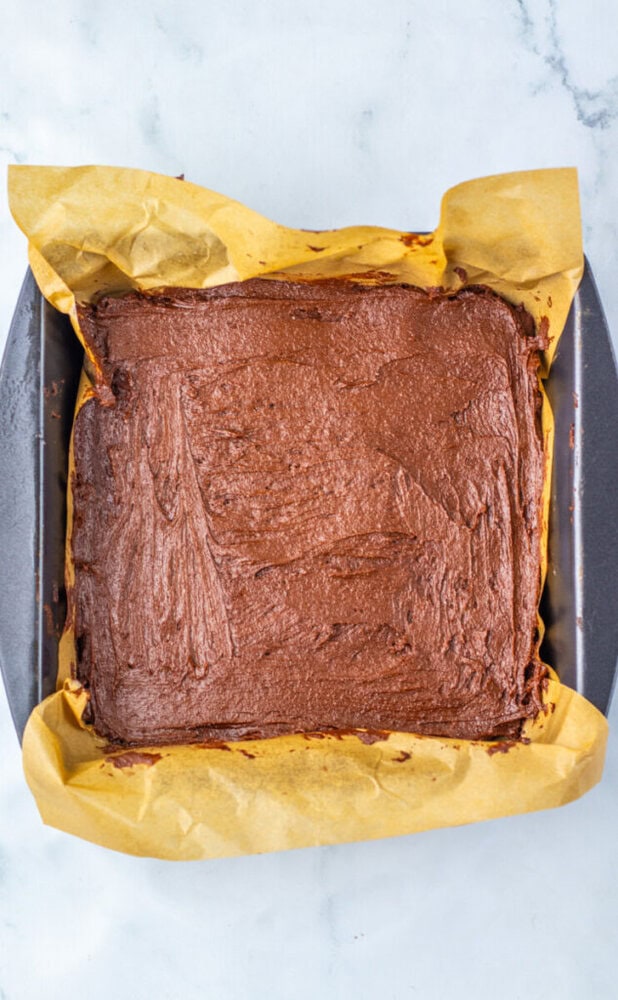 A square baking tray lined with yellow parchment paper holds unbaked chocolate brownie batter spread evenly inside.