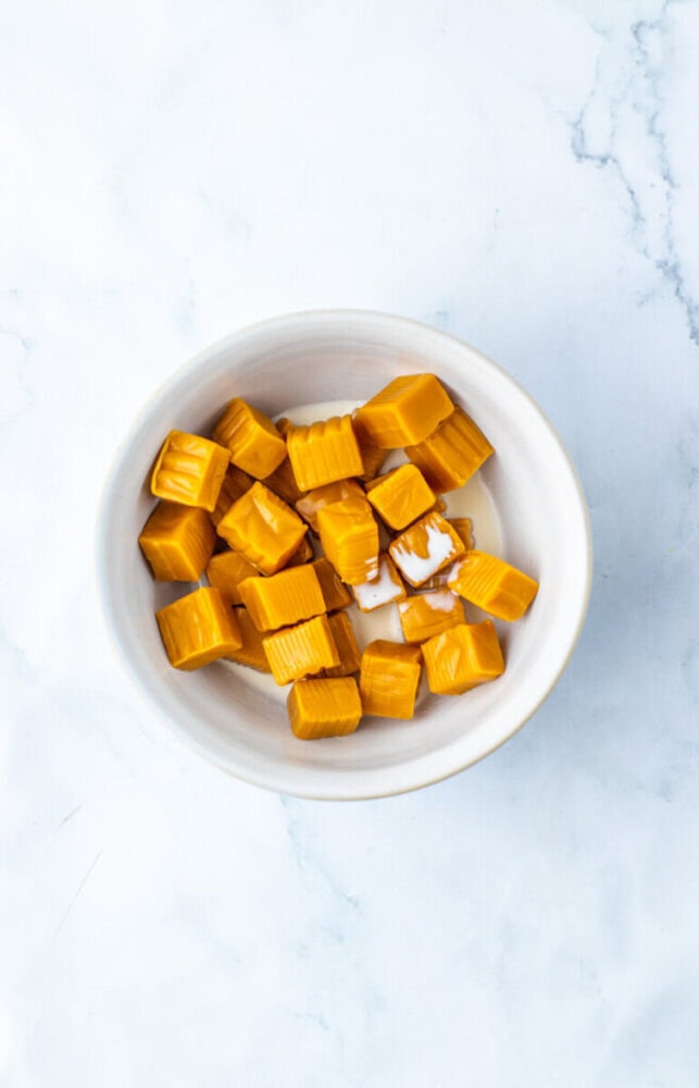 A white bowl filled with several square pieces of caramel candy on a white marble surface.