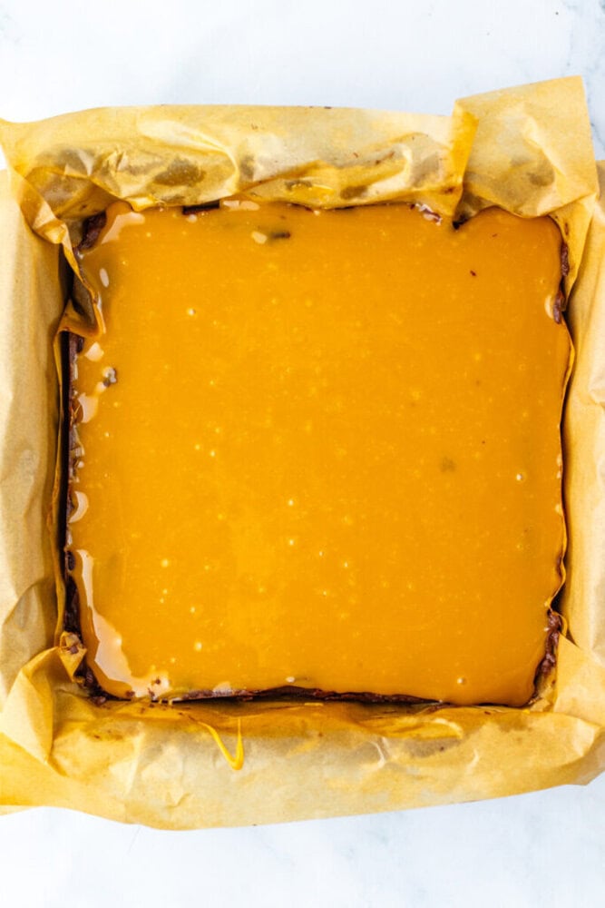A square baking pan lined with parchment paper holds a dessert topped with a smooth layer of caramel sauce.