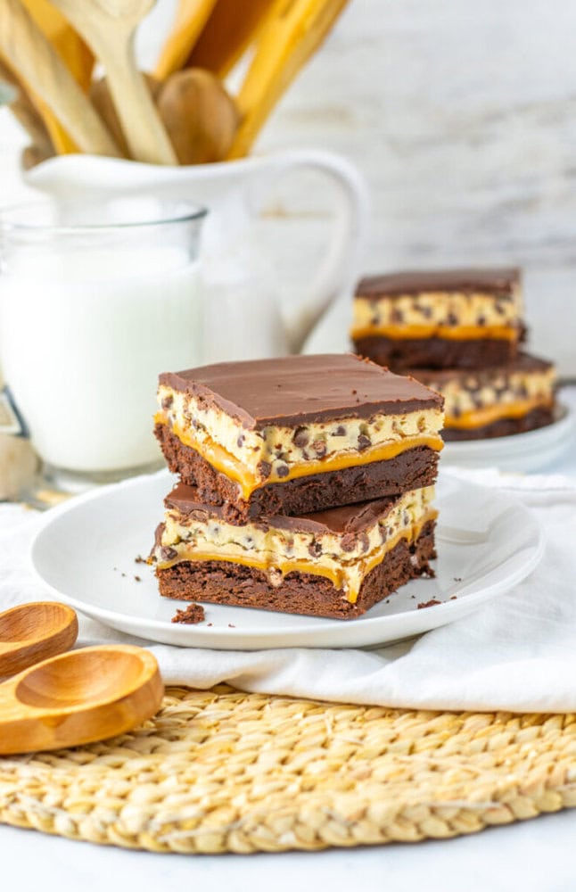 Three chocolate and cookie dough layered dessert bars are stacked on a white plate, with a glass of milk, wooden spoons, and more bars in the background.