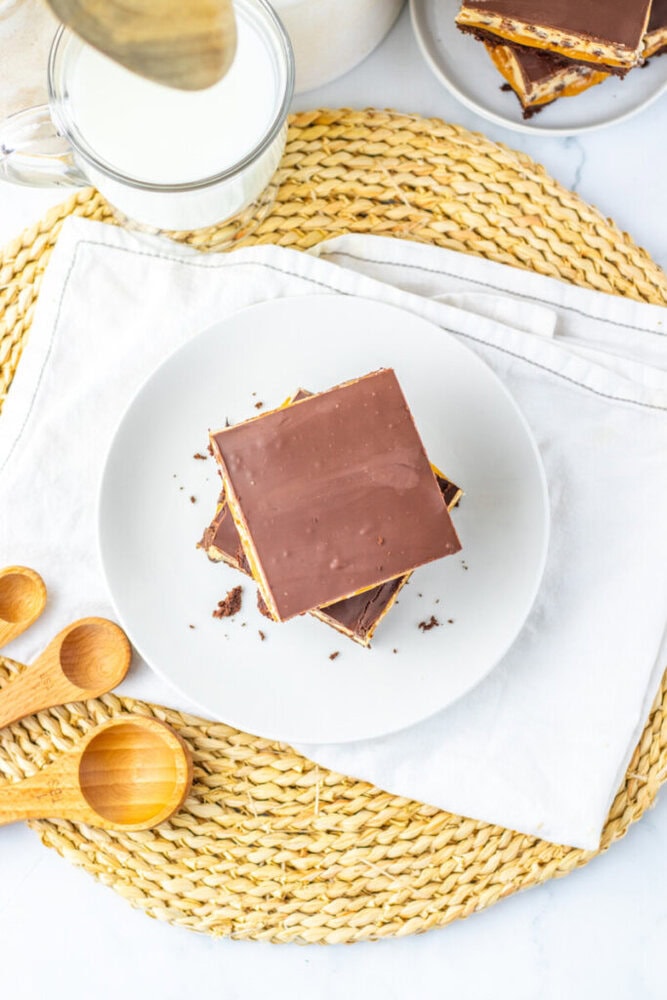 A white plate with chocolate-covered dessert bars sits on a napkin atop a woven placemat, next to a glass of milk and wooden measuring spoons.