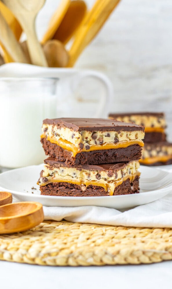 Two stacked dessert bars with brownie, cookie dough, and chocolate layers on a white plate, with a glass of milk and kitchen utensils in the background.