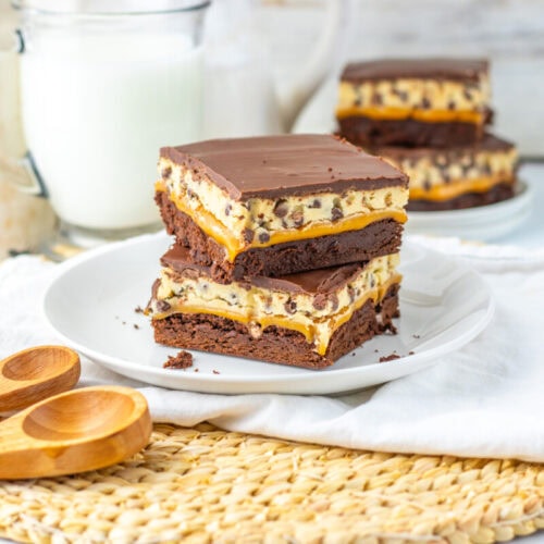 Three stacked chocolate chip cookie dough brownies on a white plate with a glass of milk and measuring spoons nearby; another serving is in the background.