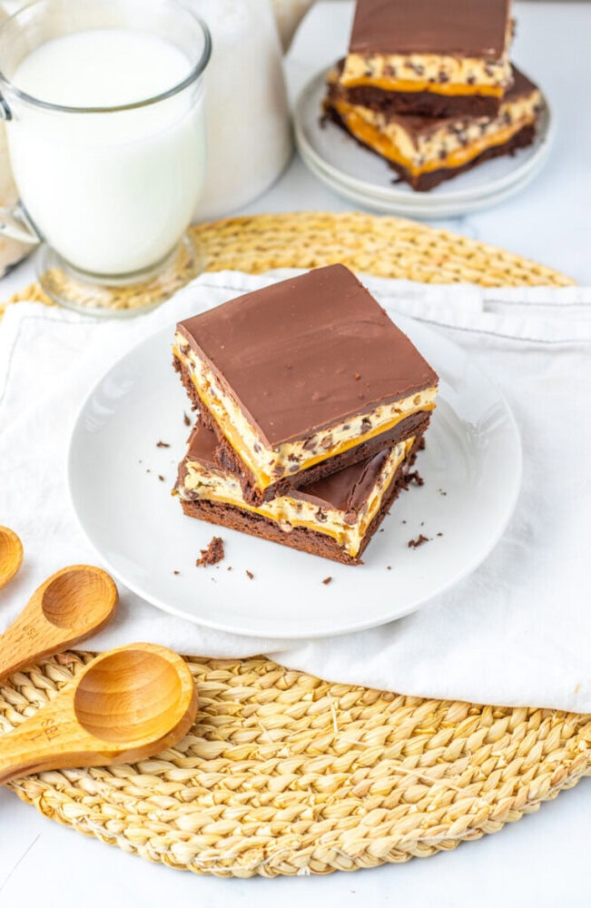 Two chocolate and cream layered dessert bars on a white plate, with a glass of milk, wooden spoons, and more bars in the background on a woven placemat.