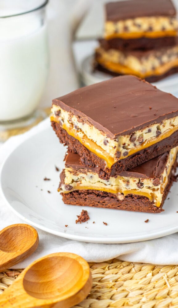Two chocolate, caramel, and cream layered dessert bars stacked on a white plate, with a glass of milk and wooden spoons nearby.